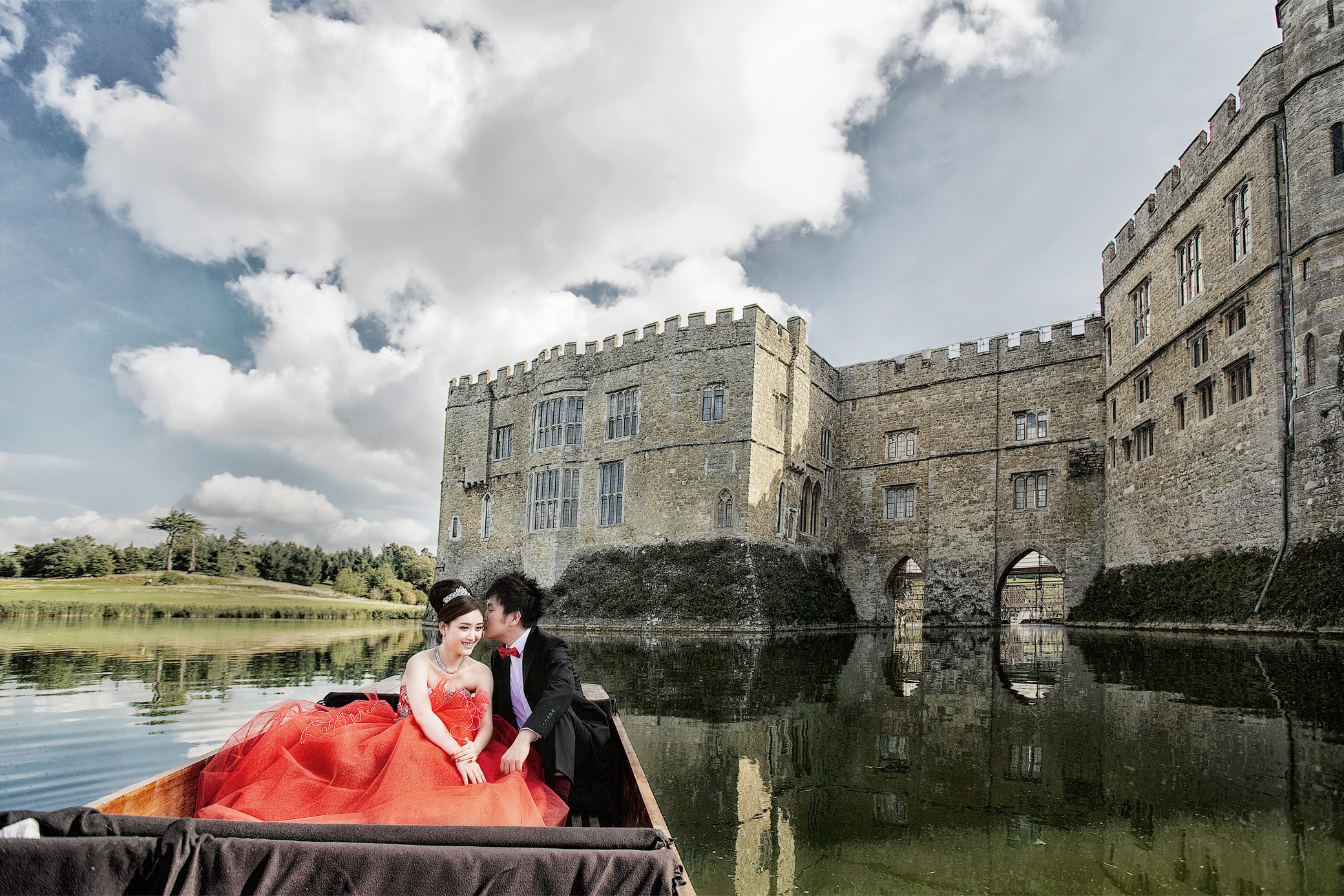 Bride in a red dress and groom in a boat on the moat below the walls of Leeds Castle