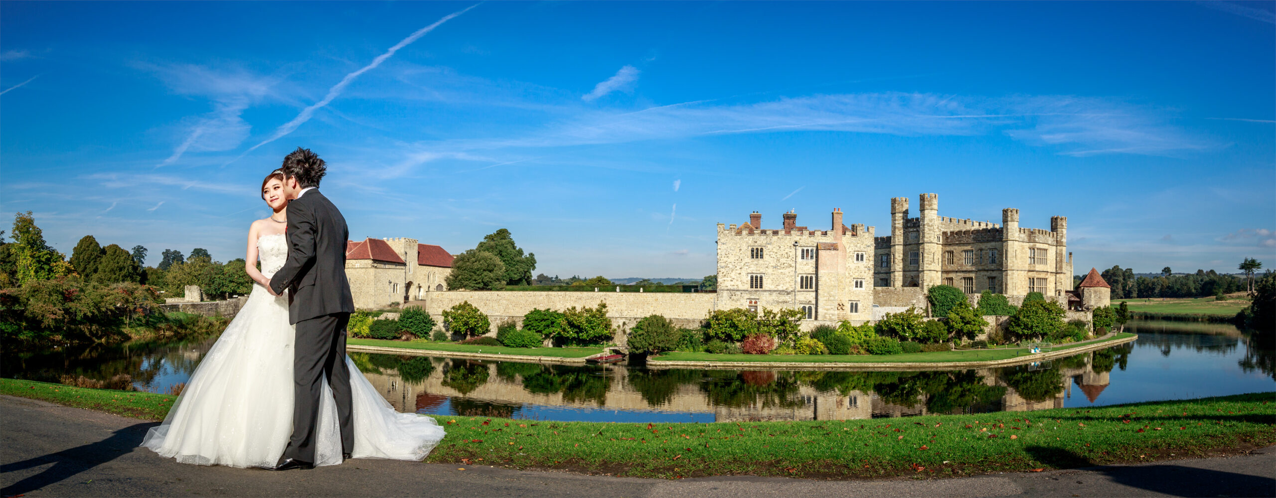 Bride and groom beside the moat with Leeds Castle across the water under a blue sky