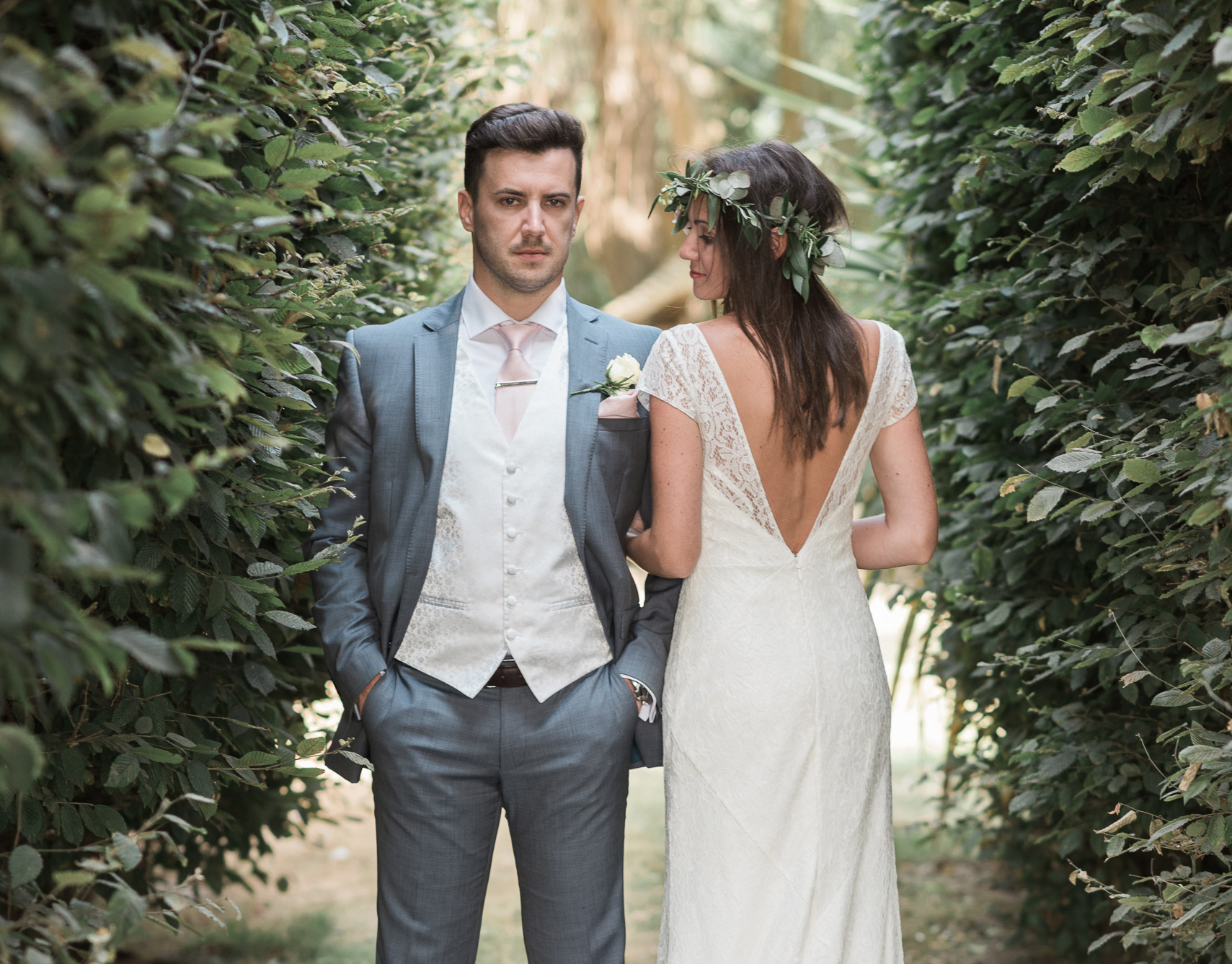 Bride with a flower crown and groom standing between tall green hedges at a Sussex garden wedding