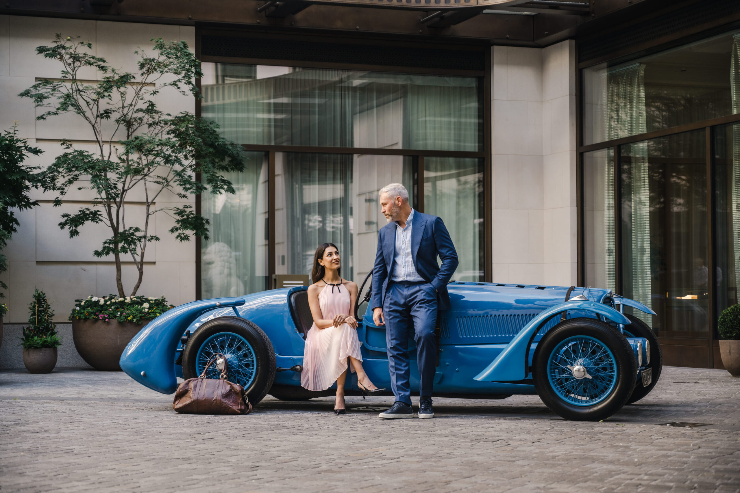 Couple sitting on the wing of a blue vintage Delahaye in the planted entrance courtyard of The Peninsula London