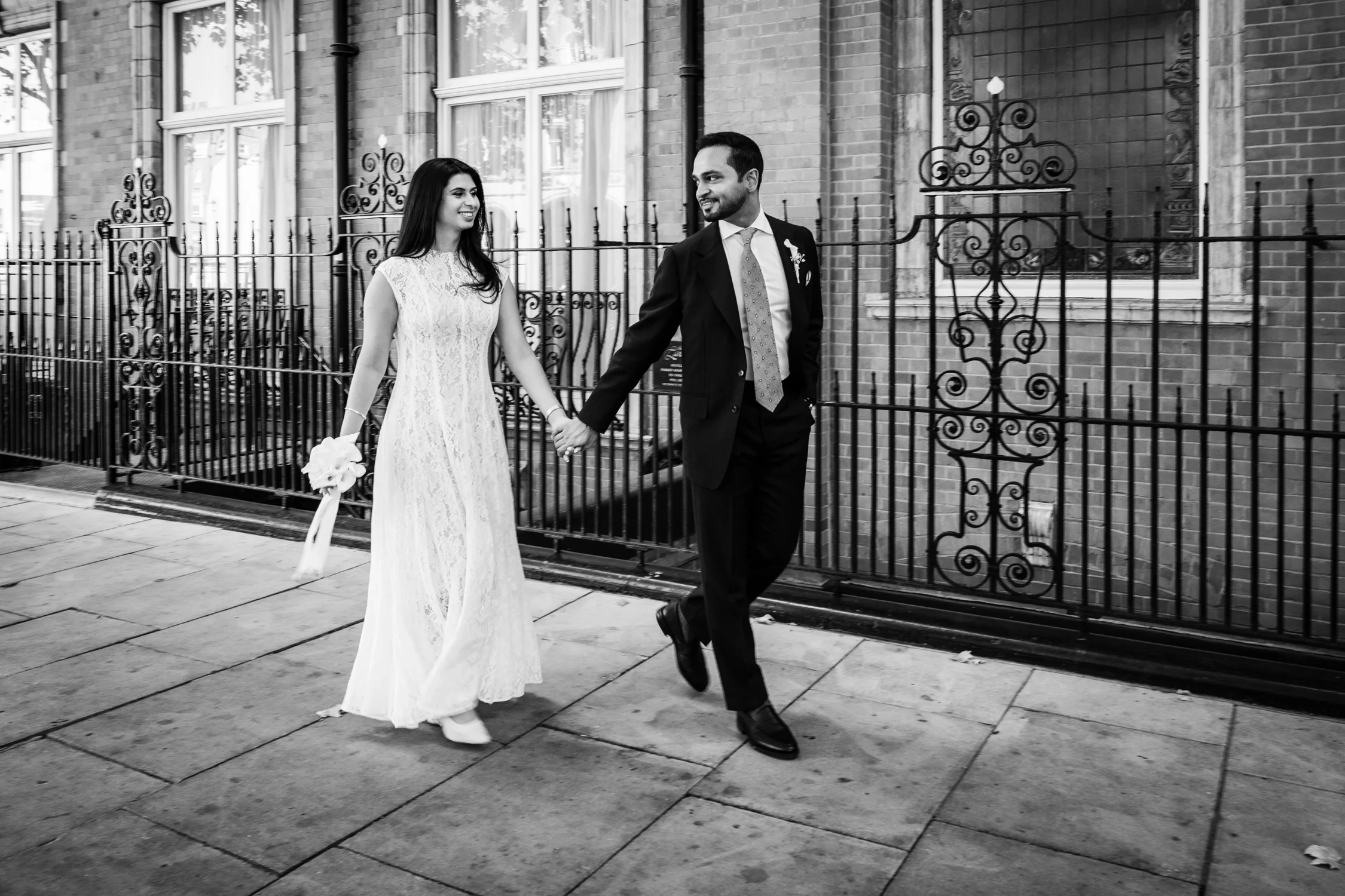 Black and white photo of the couple walking hand in hand past iron railings
