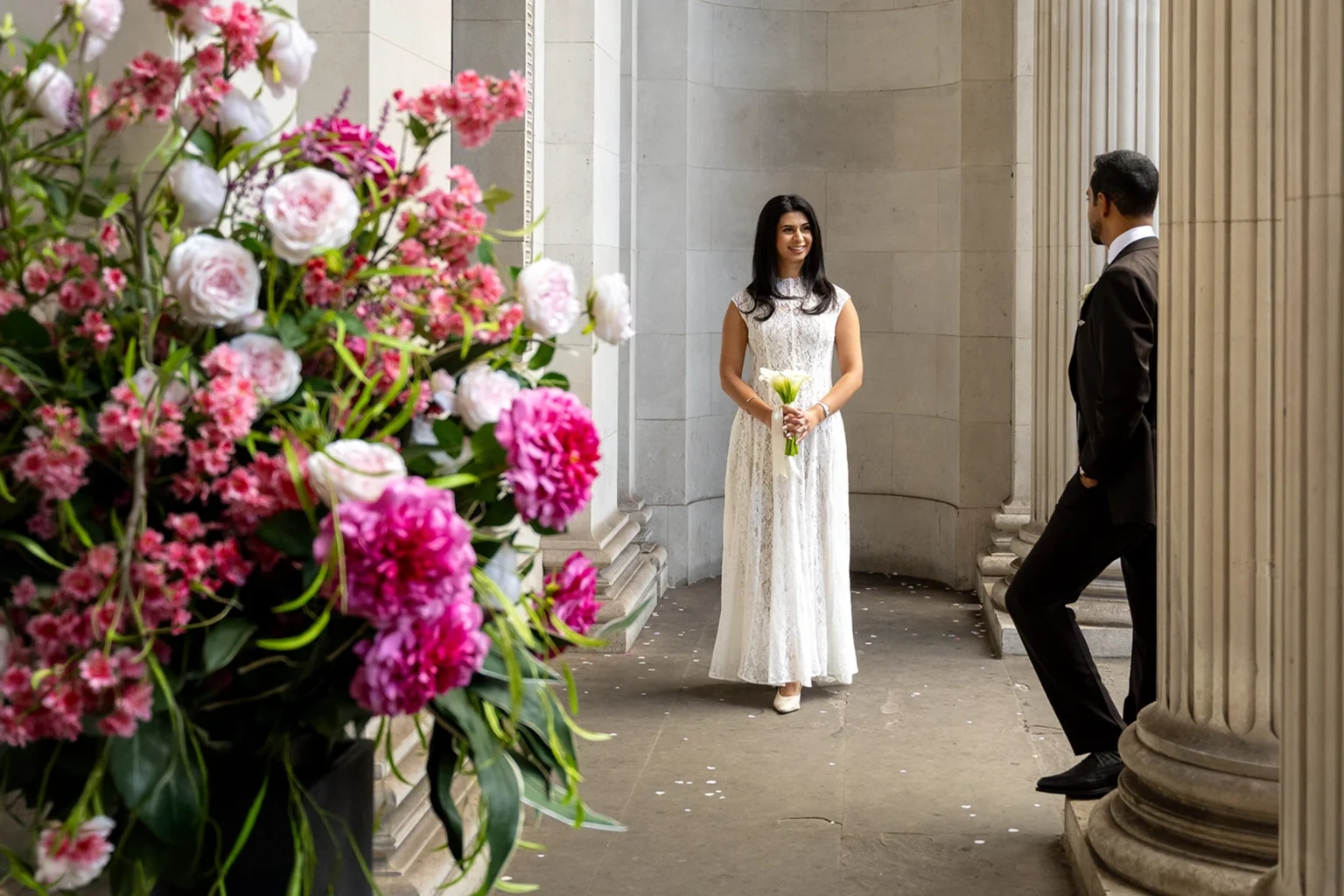 Bride waiting in the colonnade at Old Marylebone Town Hall as the groom looks back