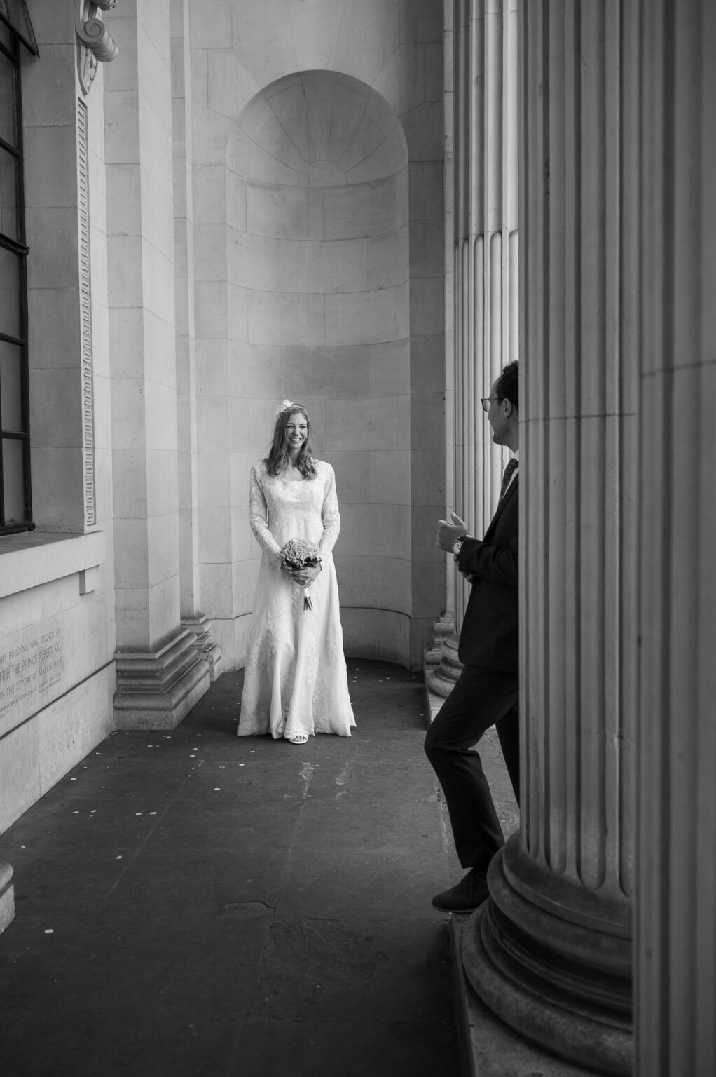 Black and white photo of the bride standing in the curved alcove at Old Marylebone Town Hall with the groom in the foreground