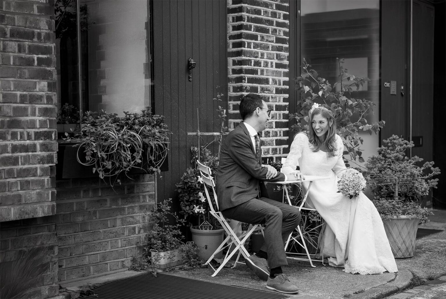 Bride and groom laughing at a bistro table on a London mews, black and white
