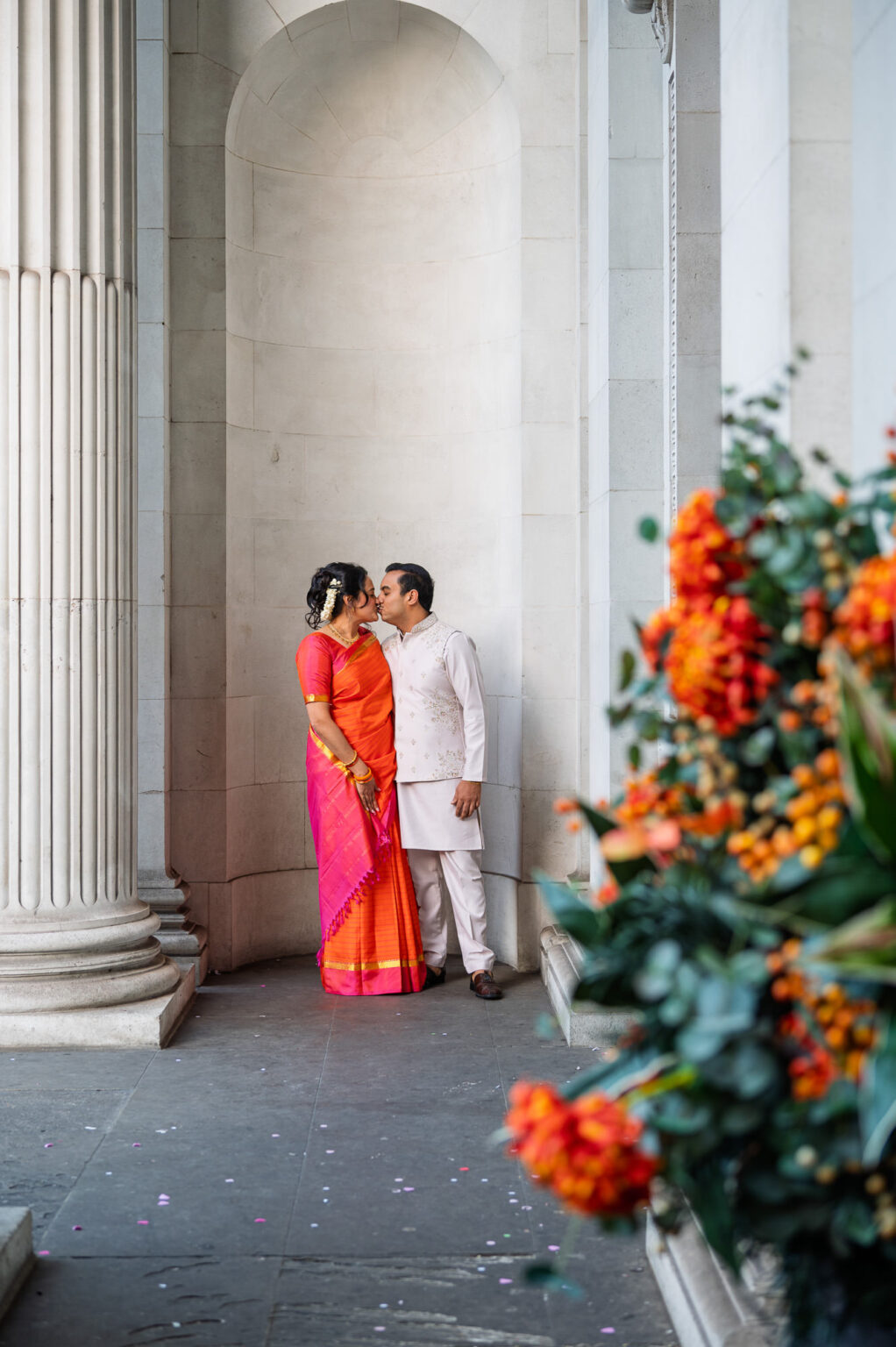 Bride and groom kissing in the arched recess at Old Marylebone Town Hall beside an orange flower display