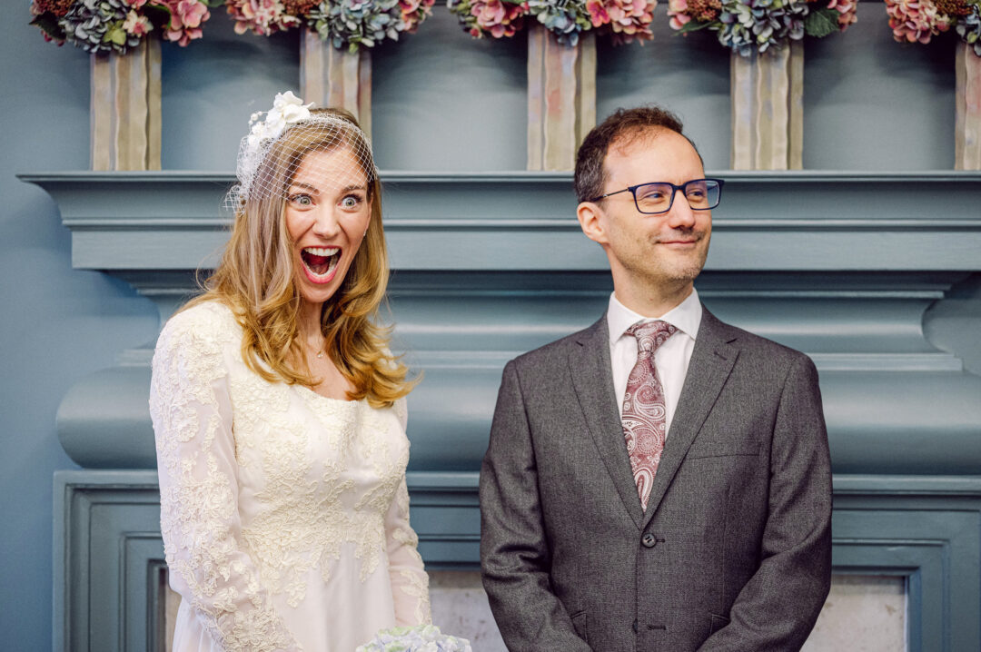 Bride with a delighted, wide-eyed expression beside her groom during their ceremony at Old Marylebone Town Hall, in front of a panelled fireplace dressed with flowers