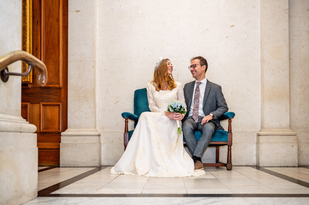 Bride and groom laughing together on a teal velvet bench in the marble entrance hall of Old Marylebone Town Hall