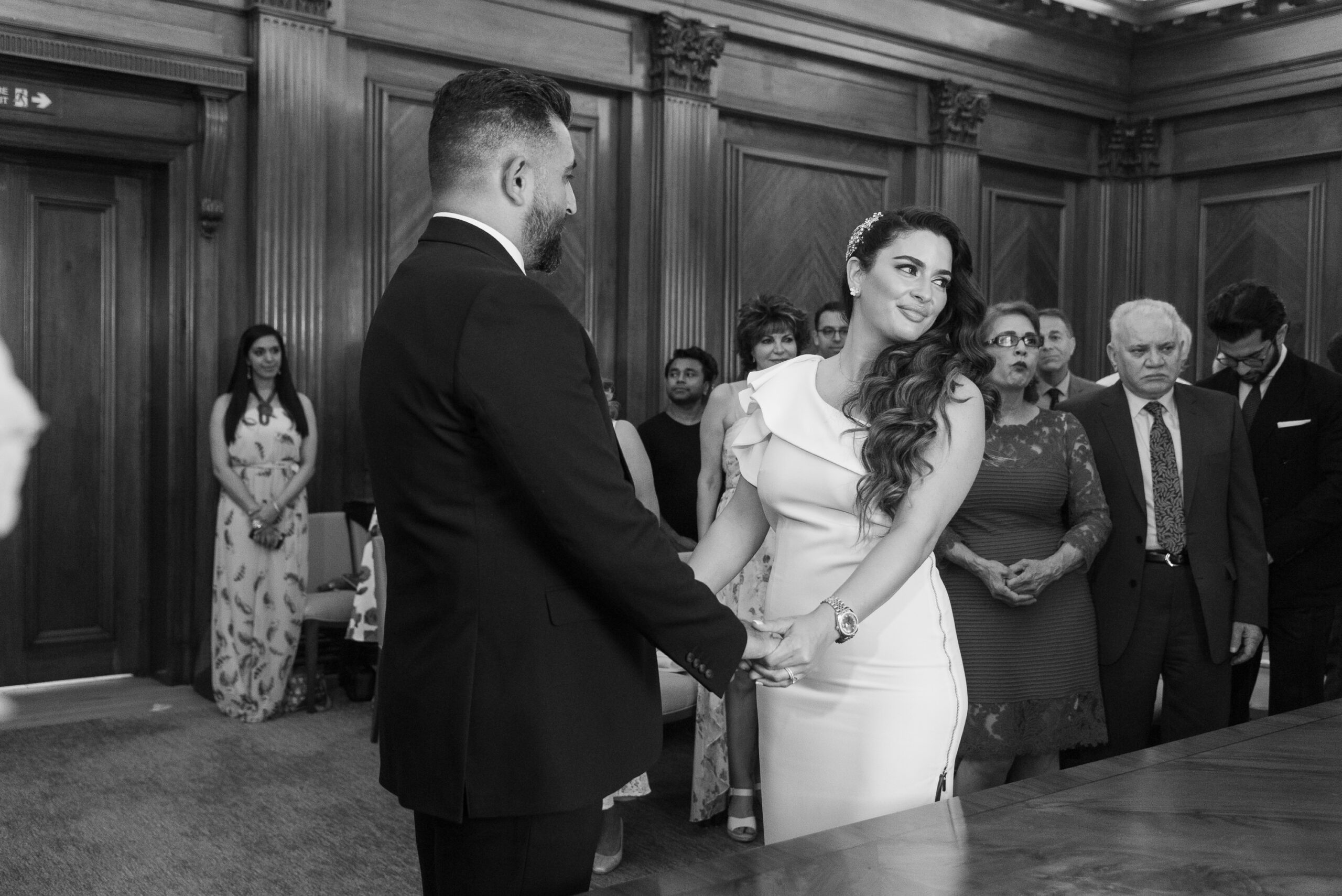 Black and white photo of Syrus and Bibi holding hands during their ceremony in the walnut-panelled Westminster Room at Old Marylebone Town Hall, guests standing behind