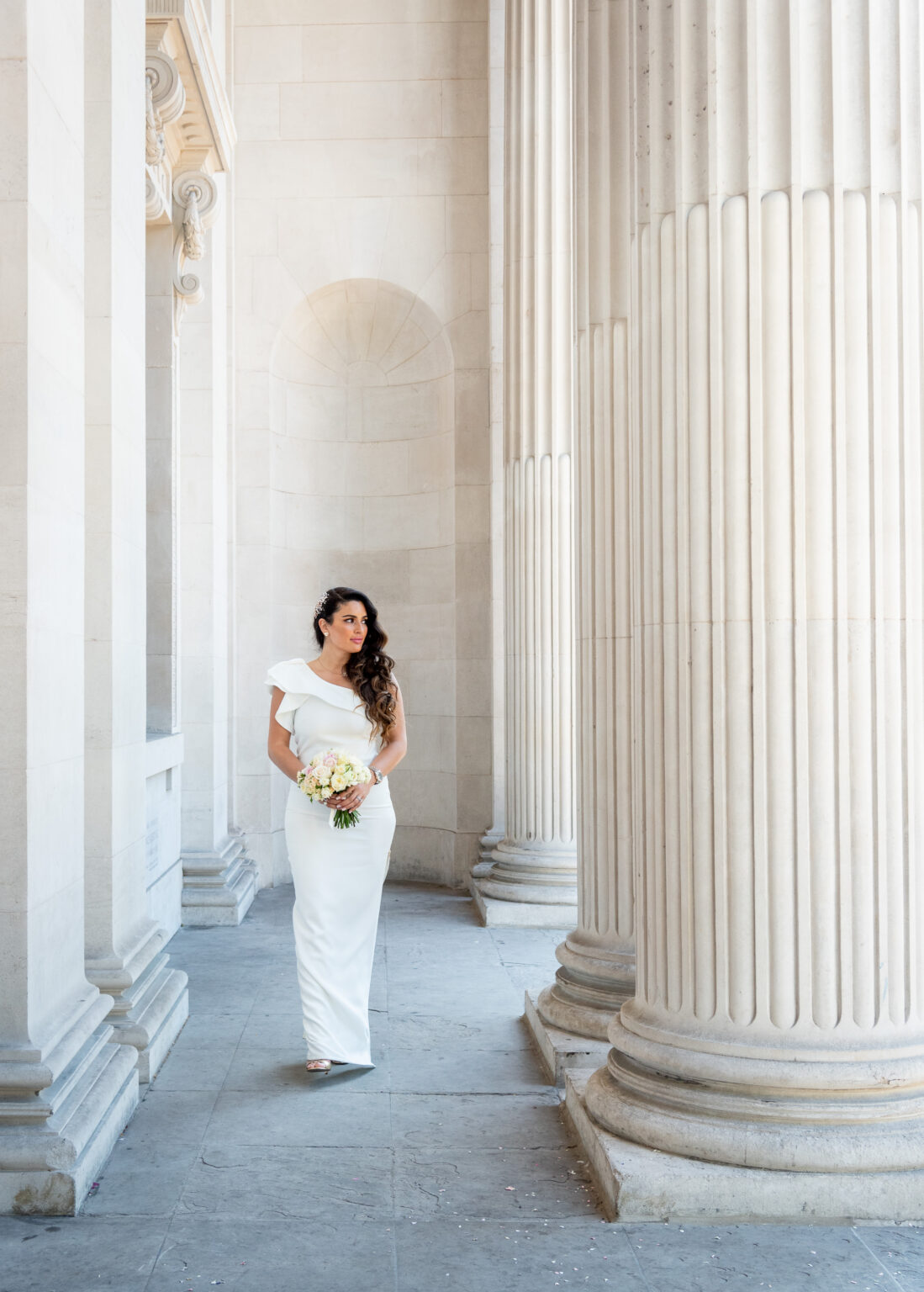 Bibi walking through the Portland stone colonnade at Old Marylebone Town Hall in a white one-shoulder dress, bouquet in hand