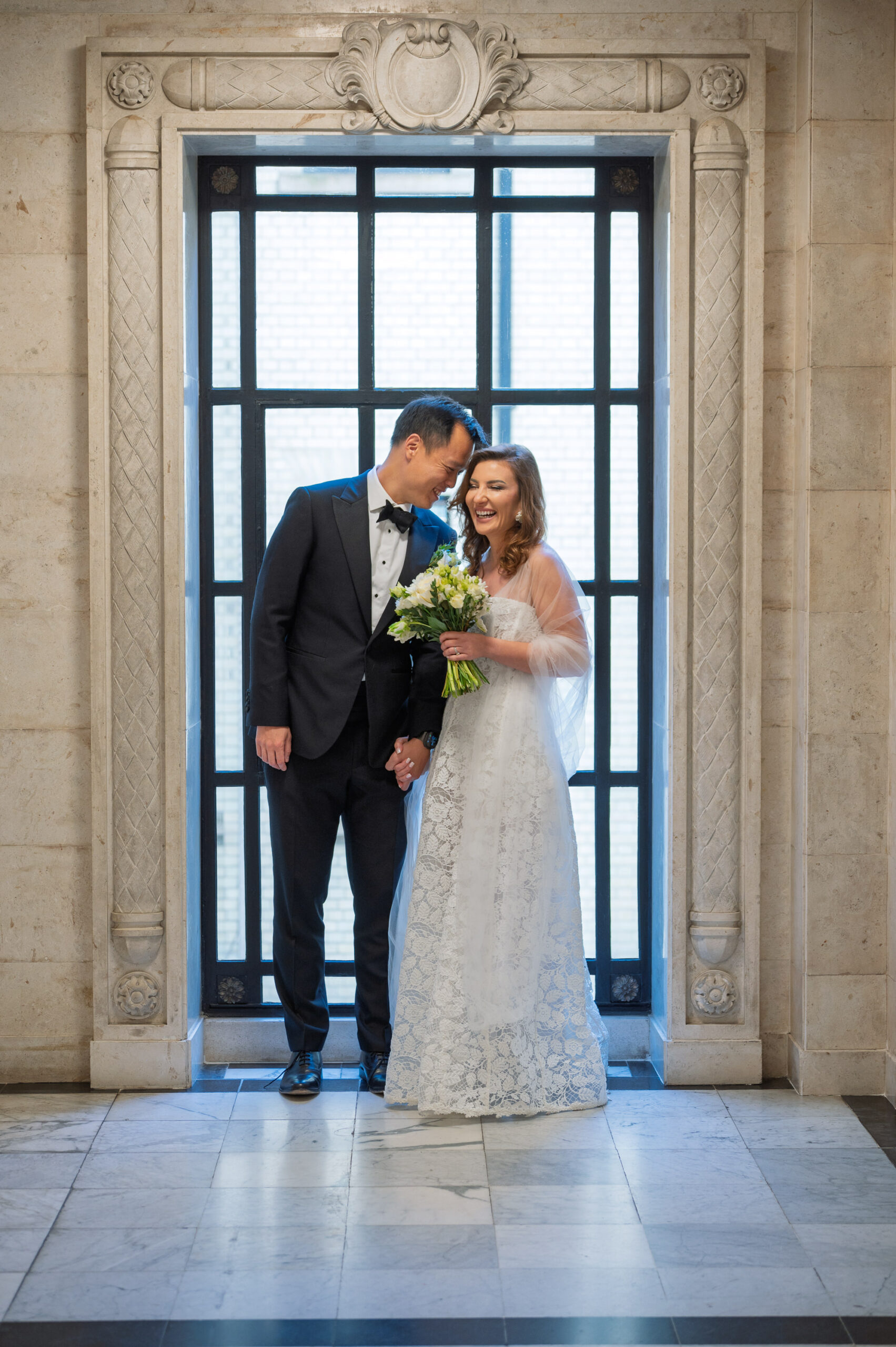 Bride and groom by the tall iron-framed window on the marble landing at Old Marylebone Town Hall