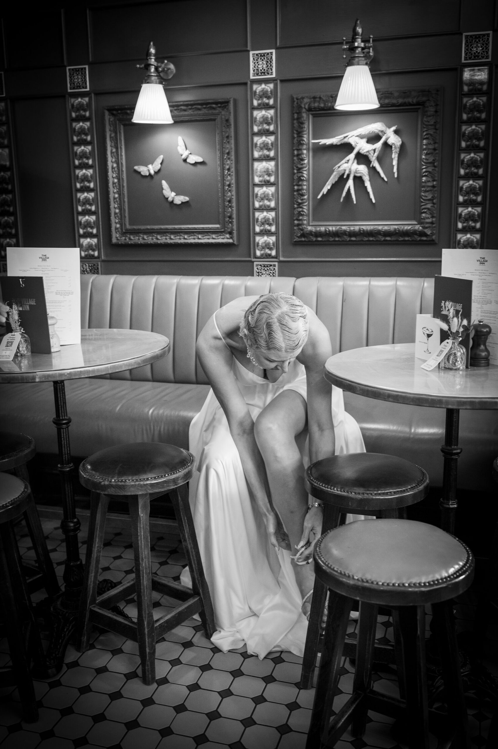Black and white photo of Nataliia fixing her shoe beside the bar stools at The Village Inn, Ealing