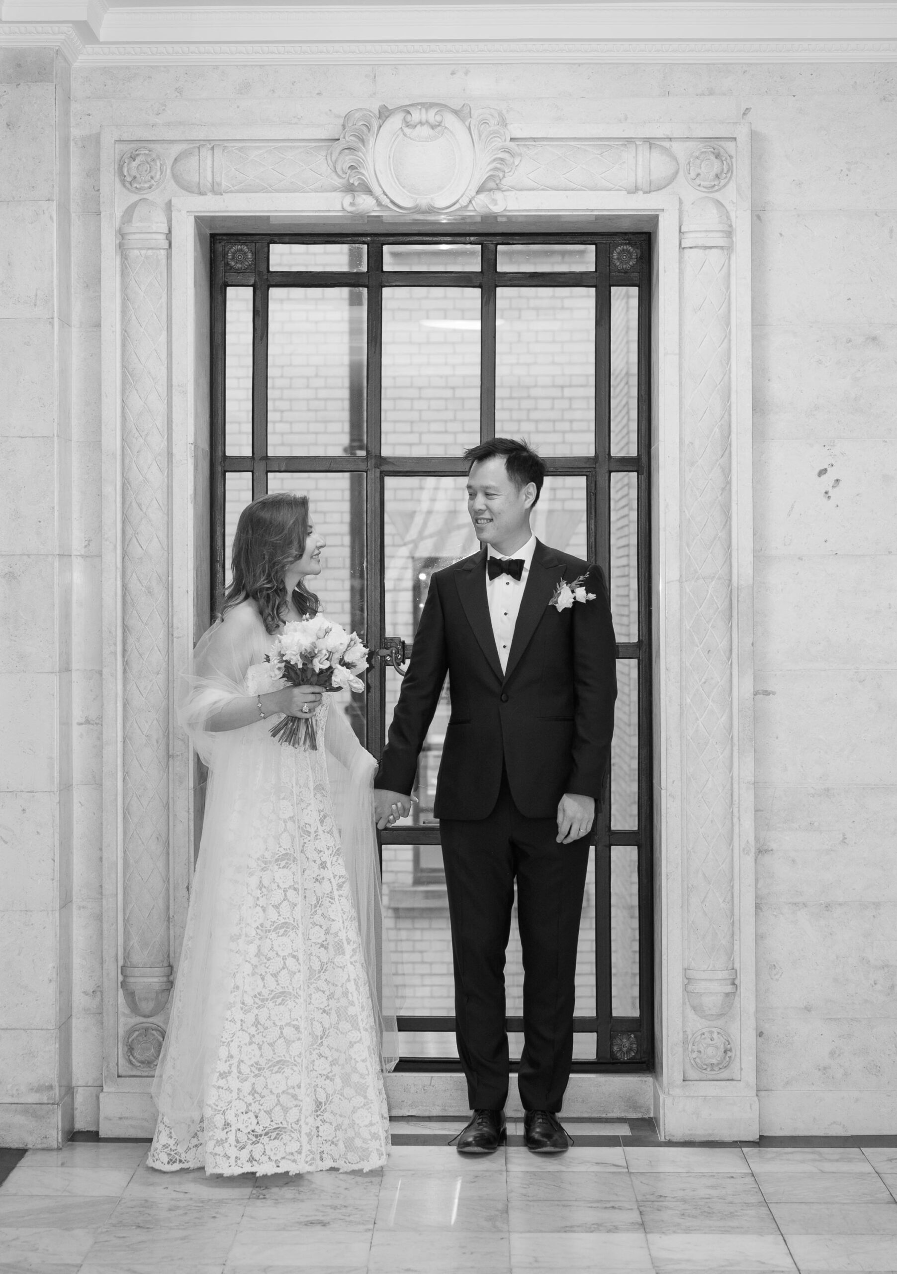 Black and white photo of Nicoleta and Alistair holding hands by the window on the marble landing at Old Marylebone Town Hall