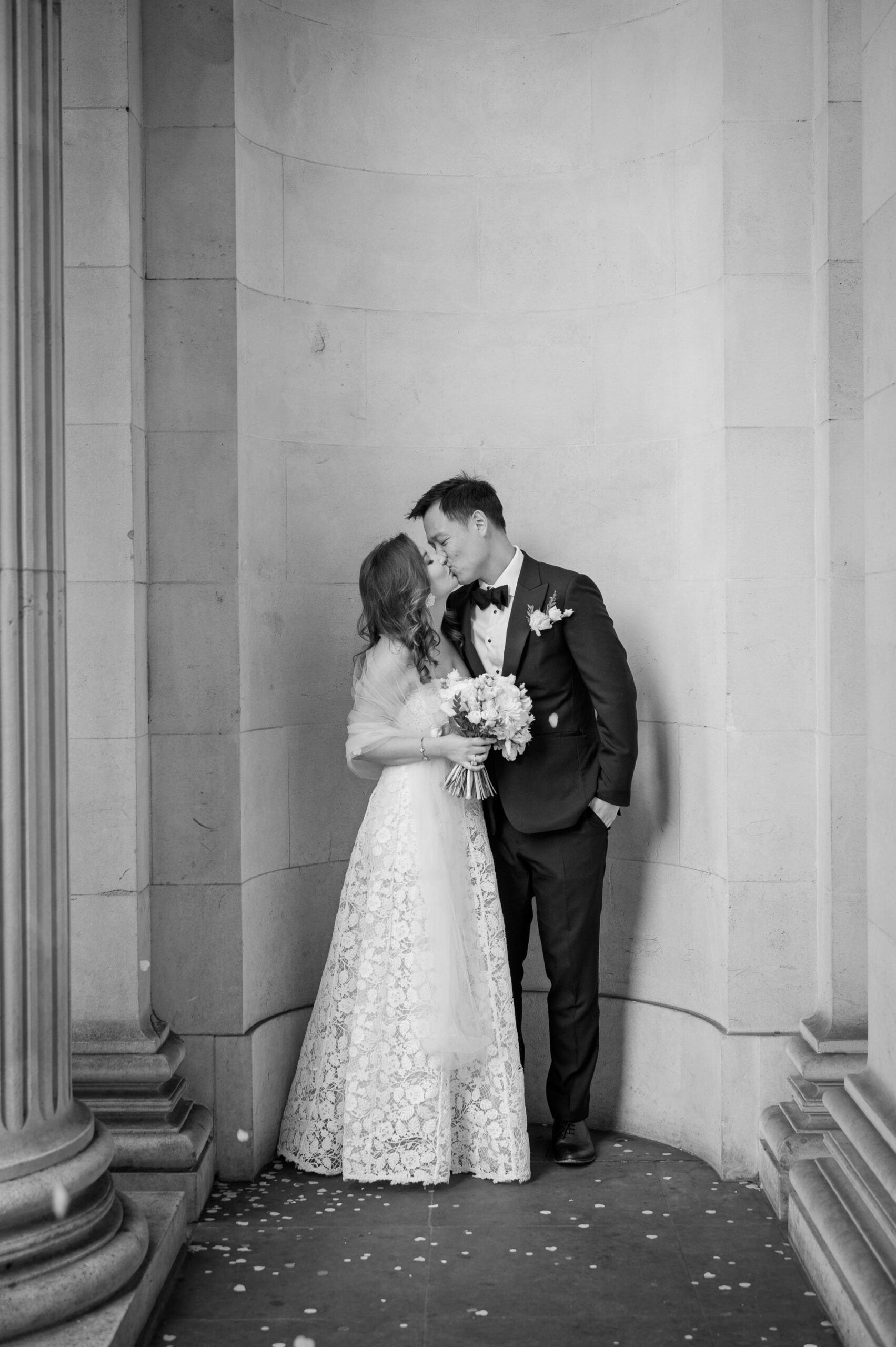 Black and white photo of the couple kissing between the stone columns outside Old Marylebone Town Hall