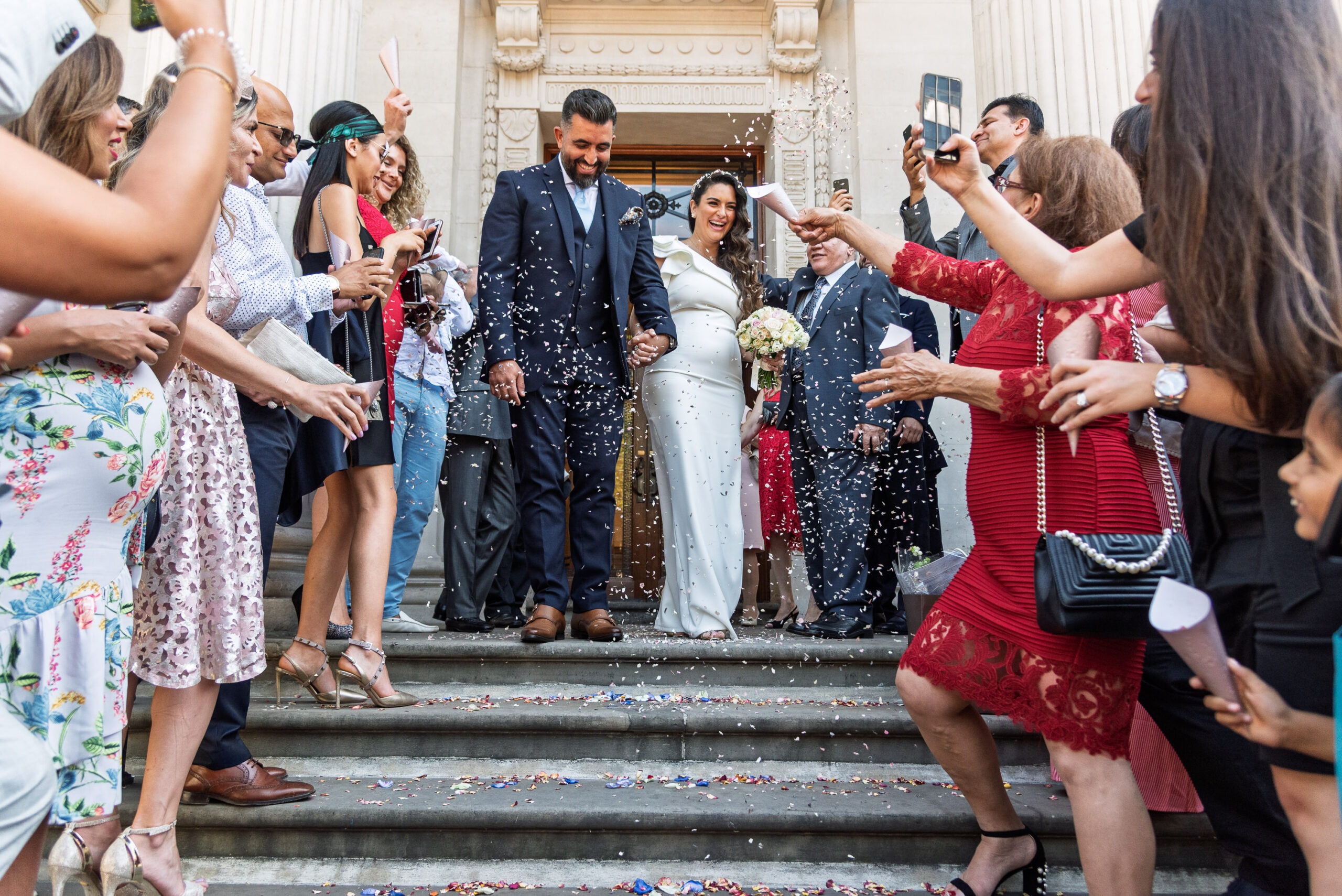 Syrus and Bibi coming down the front steps of Old Marylebone Town Hall through a shower of confetti from family and friends