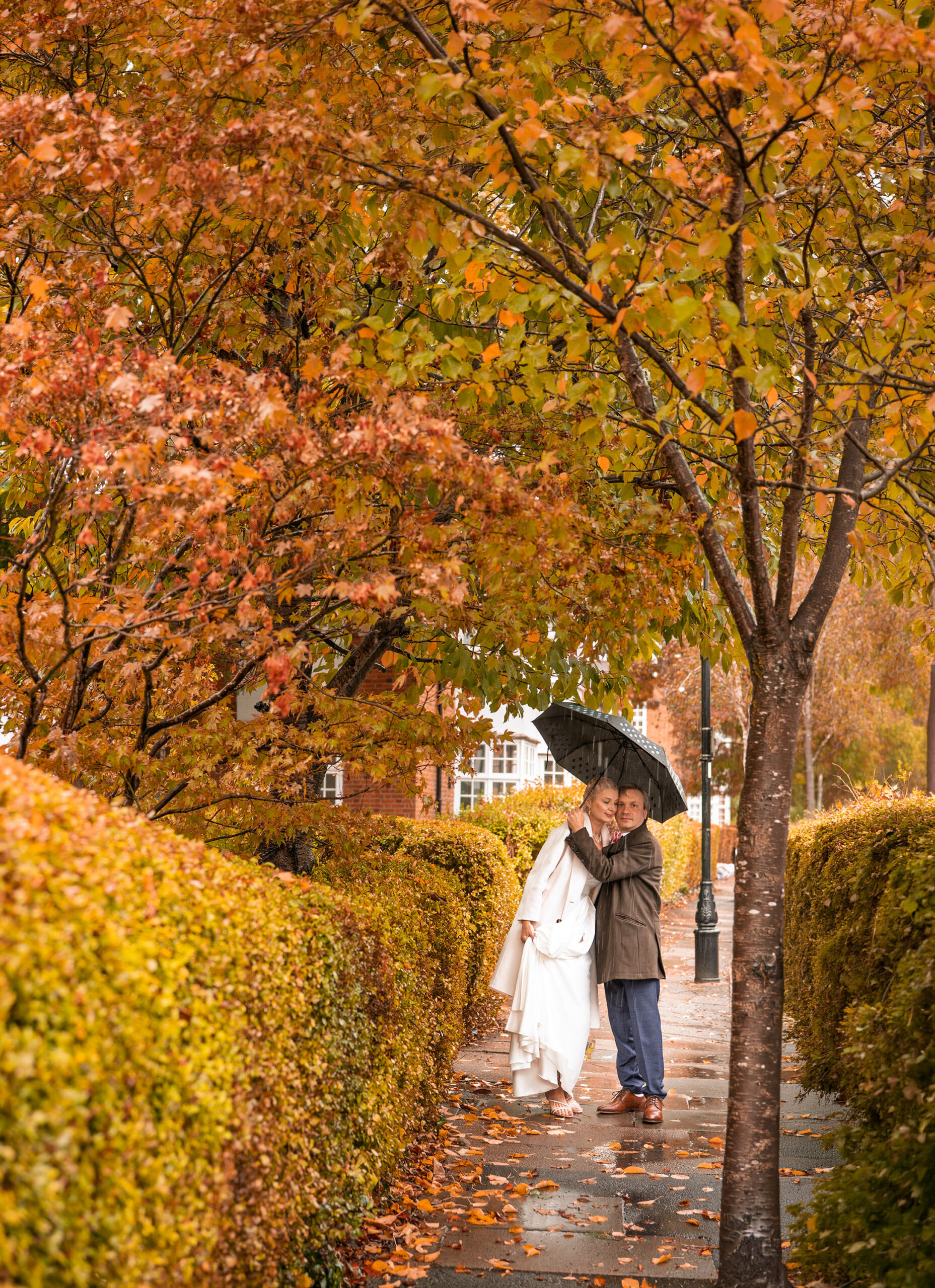 Alexei and Nataliia under an umbrella on a rainy Ealing pavement lined with orange autumn trees