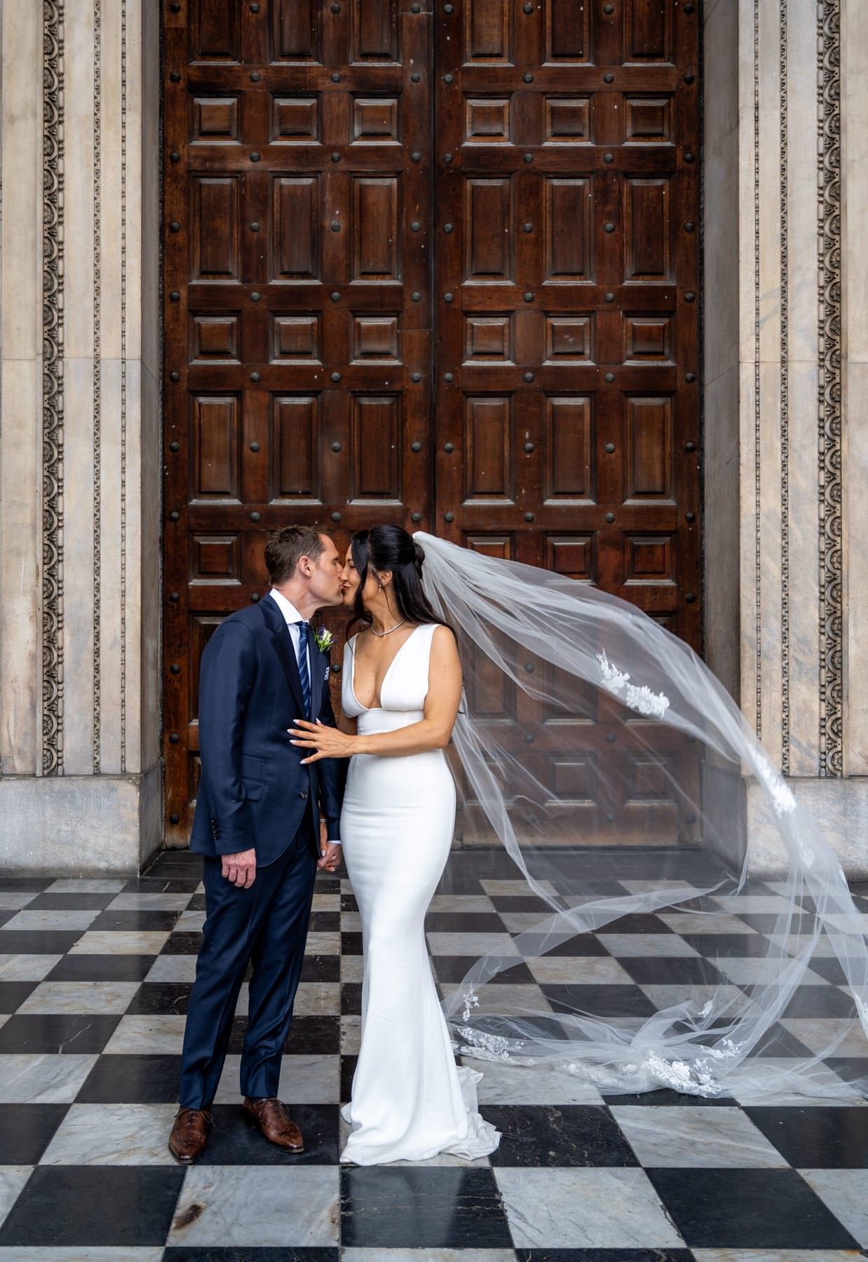 Sam and Matt kissing in front of the dark west doors at St Paul's Cathedral as the veil blows out behind them