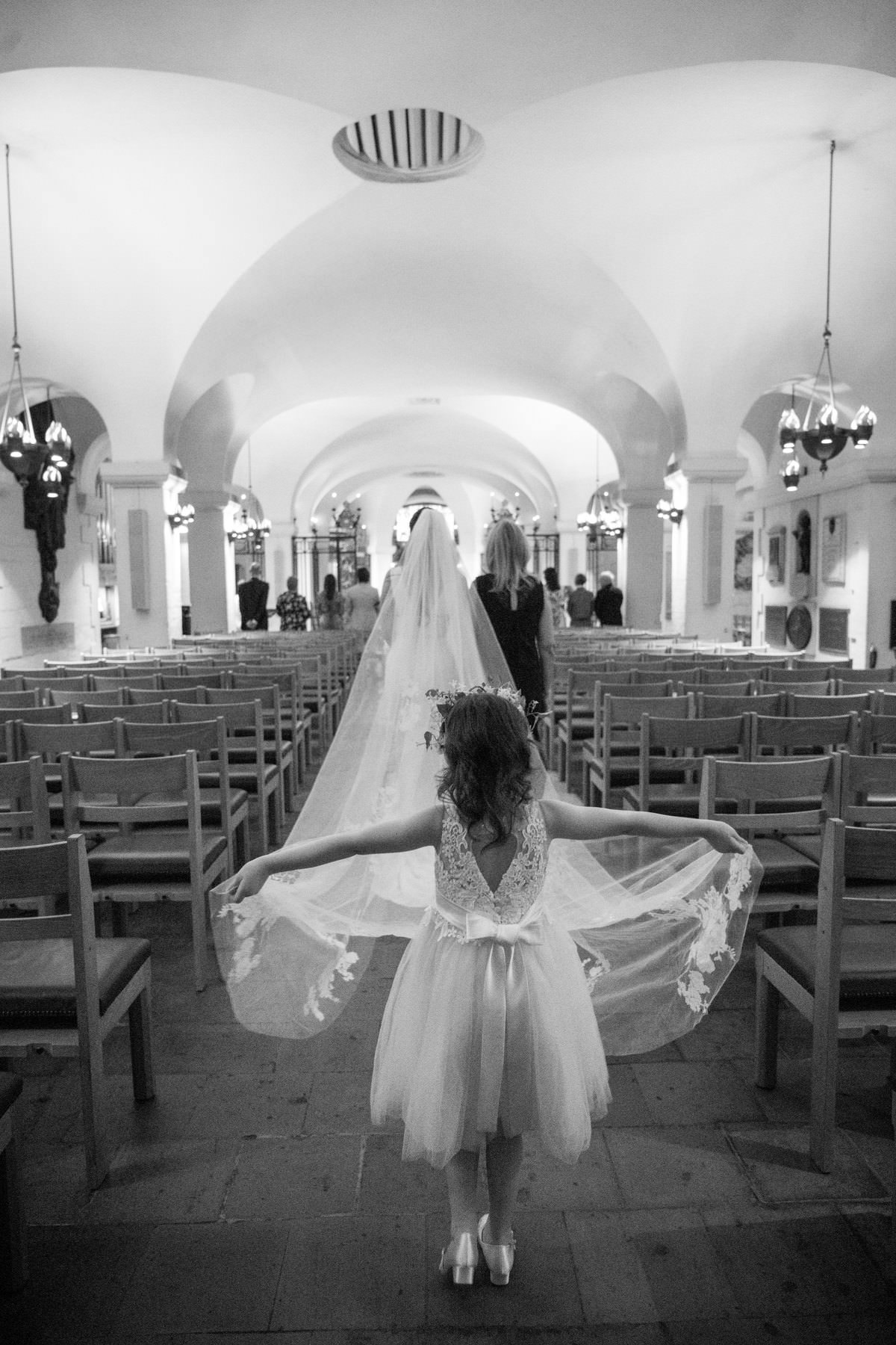 Black and white photo of the flower girl holding Sam's veil out wide as they approach the OBE Chapel in the crypt