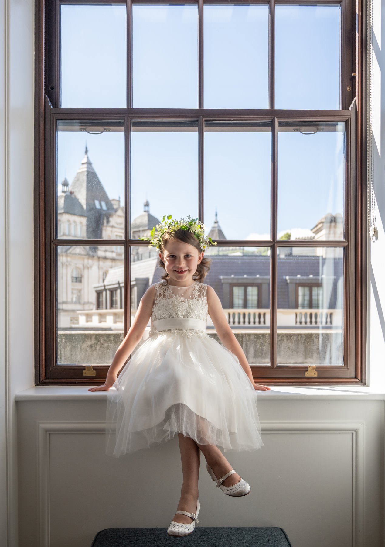 The couple's daughter in a white dress and flower crown sitting on a sash window ledge at the Corinthia hotel