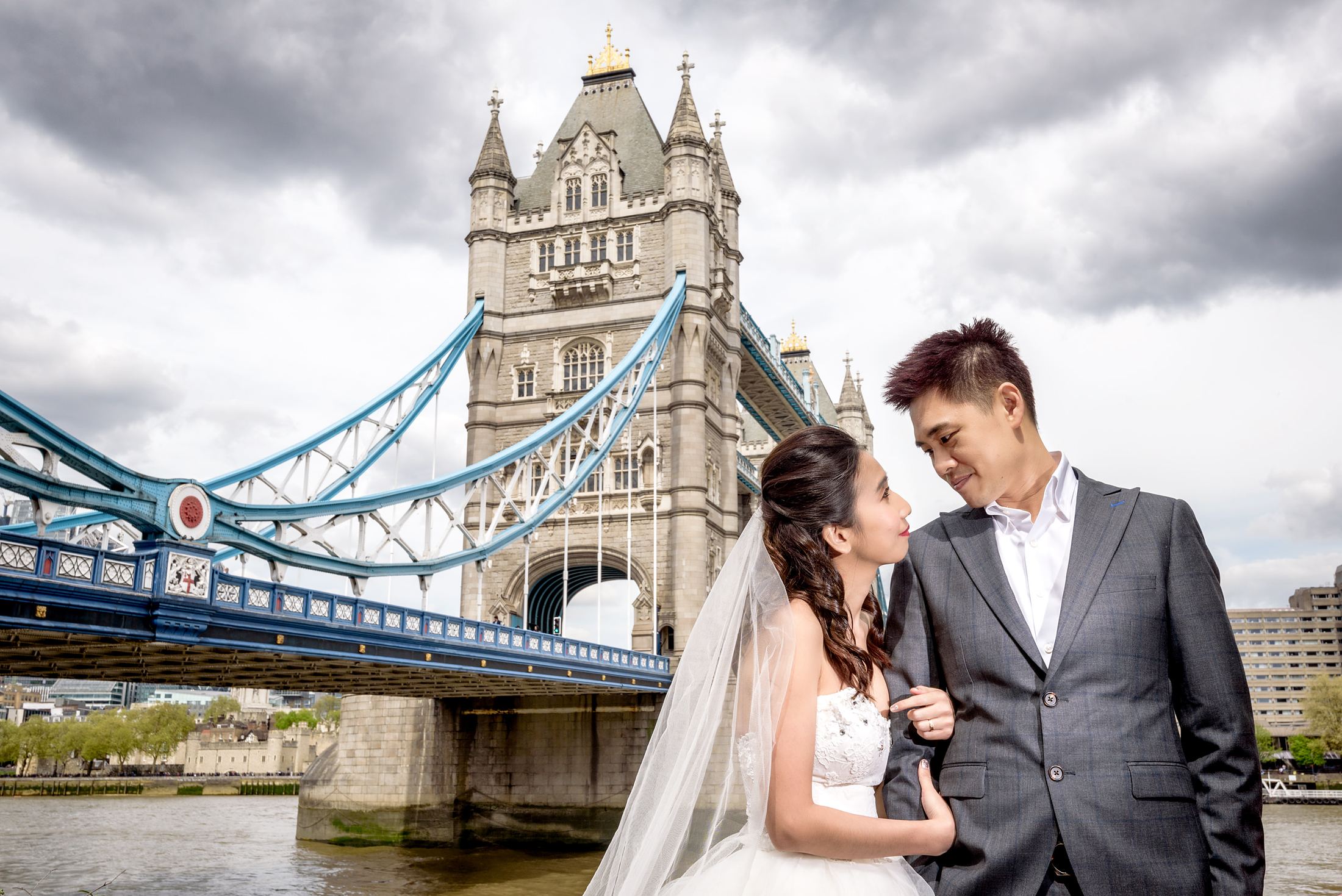 Bride in a white gown and groom in a grey suit by the Thames with Tower Bridge behind, London pre wedding photoshoot